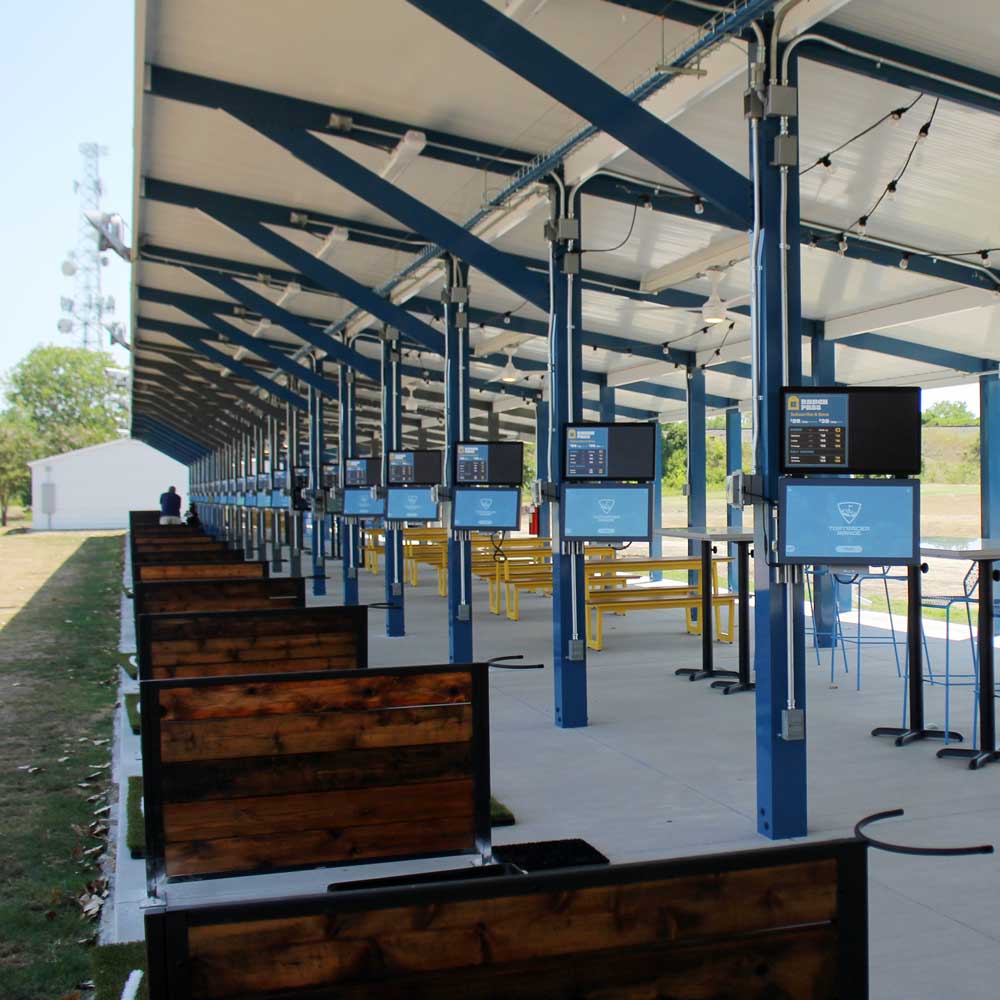 A double-row of metal pillars rising up from a concrete slab on a golf course to support a roof and create a series of open-air booths. Each booth is separated by a low wooden fence and contains a suite of sensors plus a pair of video monitors.