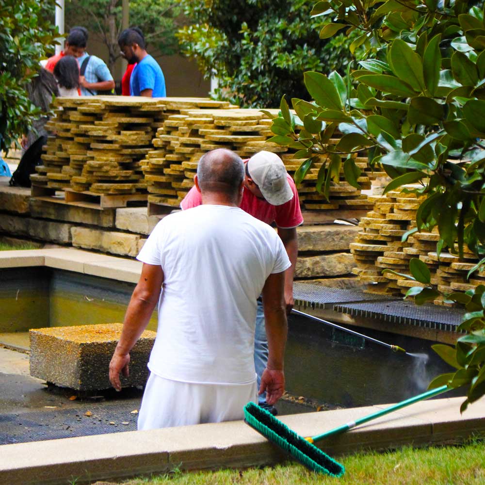 Seen through the leaves of magnolia trees, two workers stand in the corner of a drained shallow rectangular pool, powerwashing the walls and moving grills. Stacked along one edge of the pool are the flat rocks which normally form its patterned floor. Behind them, up at ground level, is a gathering of students.