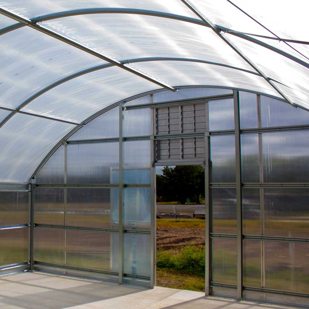 The inside of a glass and steel arched-roof greenhouse. It is empty of plants for now, but filled with light. An open door at the far end looks out onto a small field.