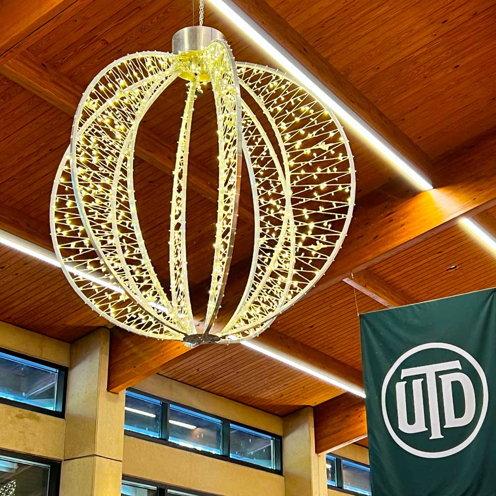 From a polished wooden ceiling hangs a green banner bearing the UTD monogram and large holiday ornament made from loops of silver metal wrapped in strings of golden lights.