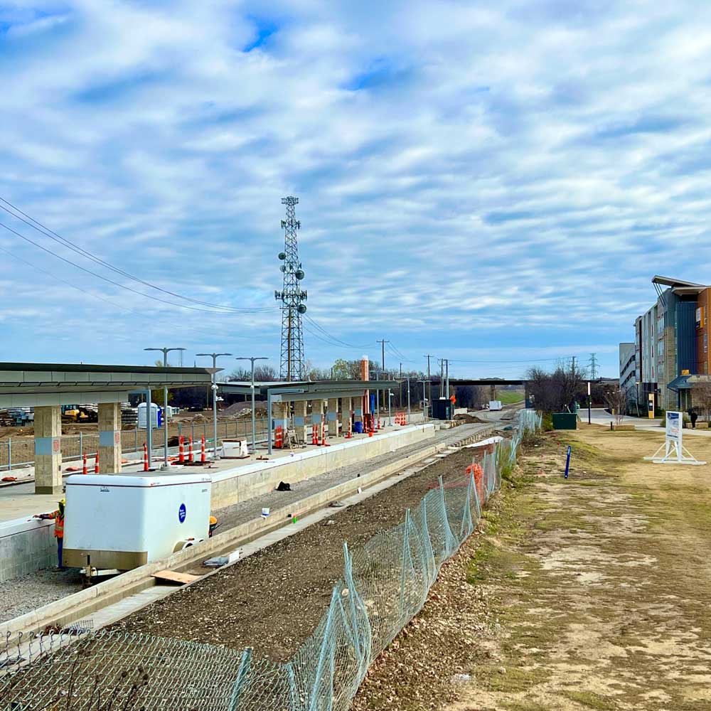 The construction site of a commuter rail transit station, with a strip of bare land running towards the horizon that passes a nearby apartment complex and goes under a bridge. Several support columns on the station bear the UTD logo.