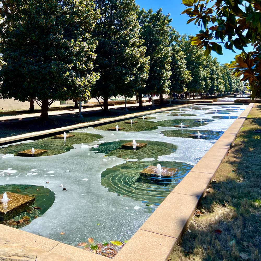 A long row of rectangular pools extends into the distance between two rows of magnolia trees. A circle of open water surrounds each bubbling fountain in the otherwise solid sheet of ice that covers every pool.