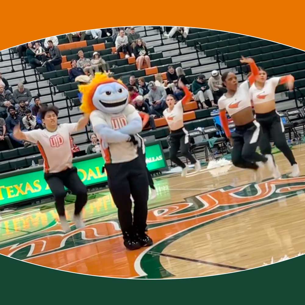 Temoc, the University mascot built like an anthropomorphic comet, and several UT Dallas Power Dancers leaping in the air during a cheer routine in the middle of a basketball court.