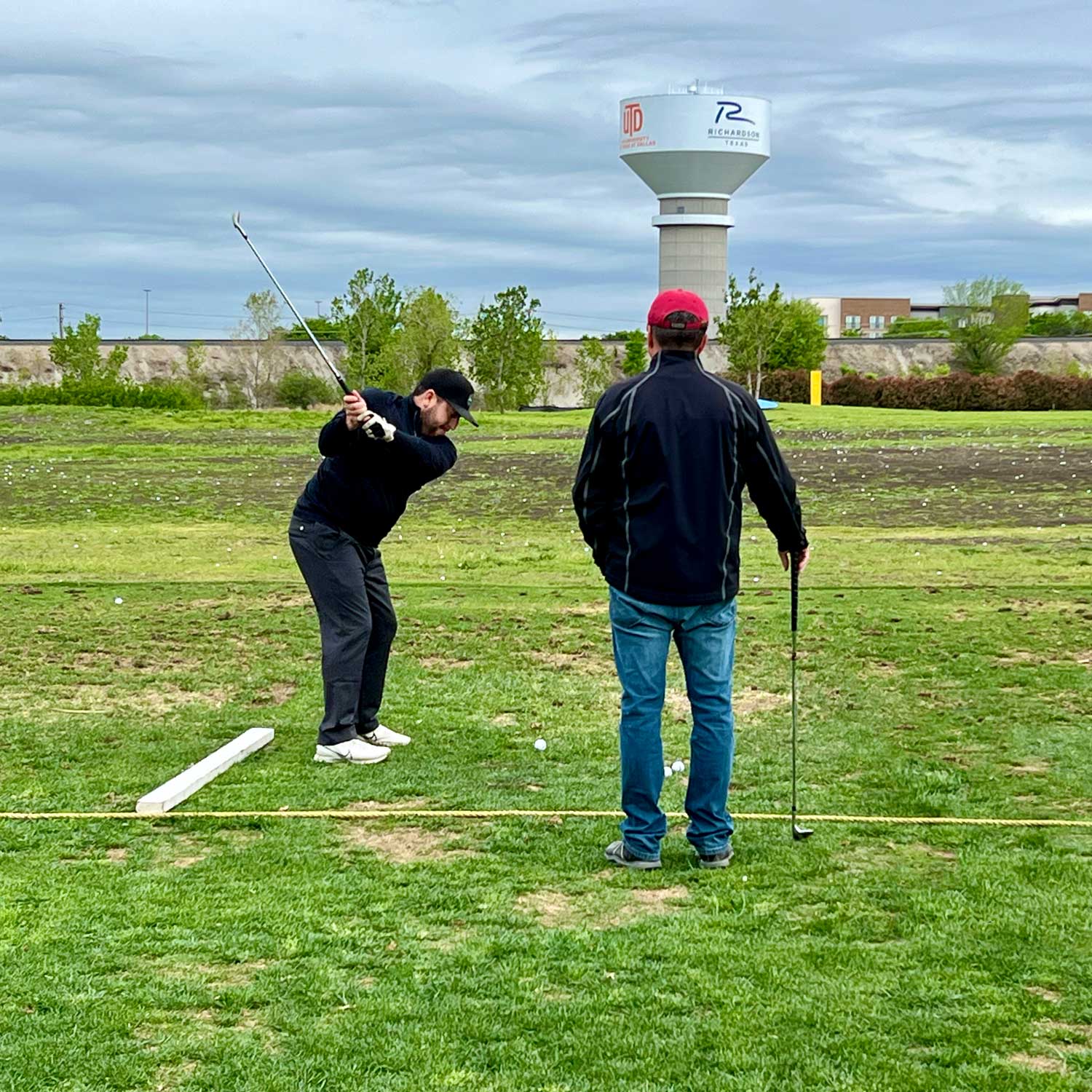 A student practices his swing under the observation of a golf instructor on the main driving range of Golf Ranch Richardson. In the distance beyond a hill, a water tower bearing the UTD and City of Richardson logos rises into a cloudy sky.