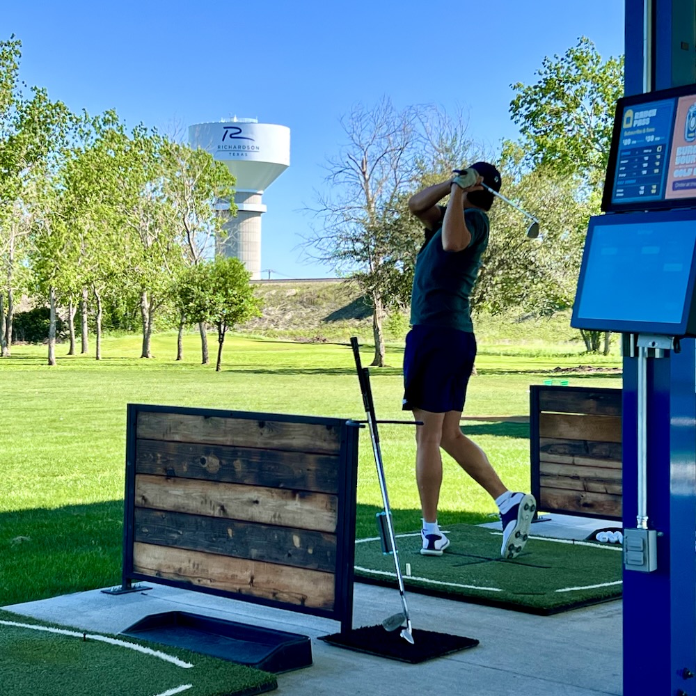 A student golfer tees off on a sunny afternoon from a practice tee filled with monitoring equipment, out into a tree-bordered grassy lawn, with the City of Richardson water tower in the distance.
