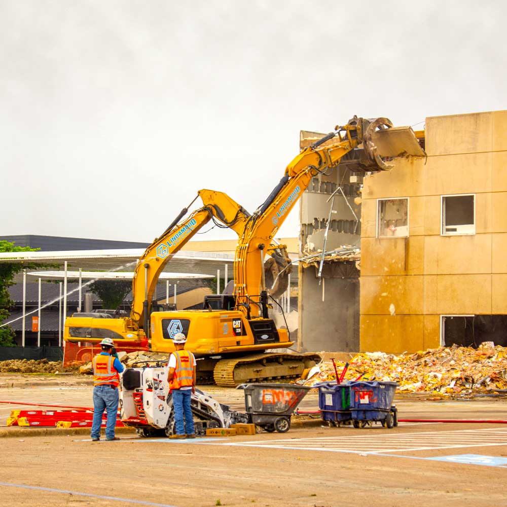A pair of demolition excavators tear apart the walls of a stone structure as two construction workers in safety gear look on.
