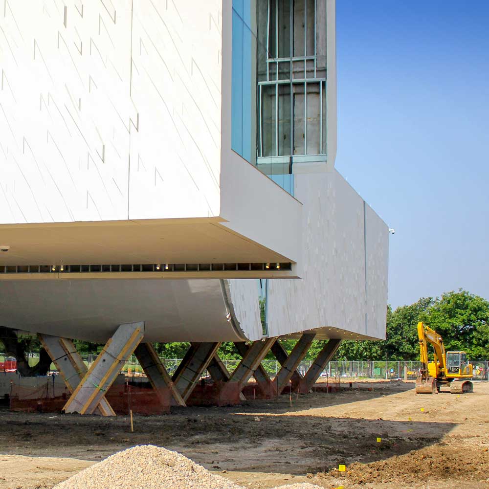 A modern building covered in textured concrete panels rises above a construction site on v-shaped supports. A piece of construction equipment stands parked nearby.