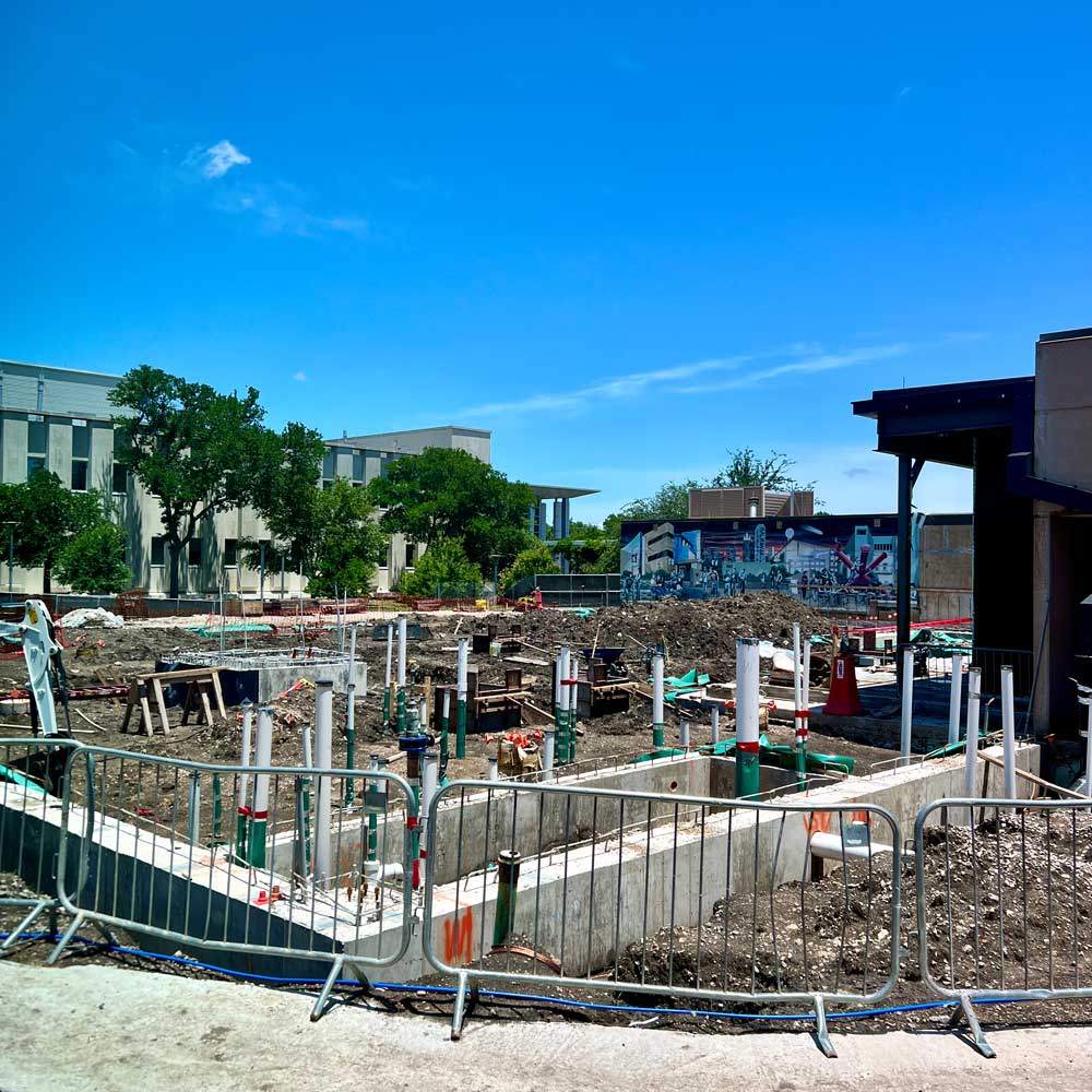 A fenced-off construction site in the middle of a college campus, from which various pipes rise out of the ground.