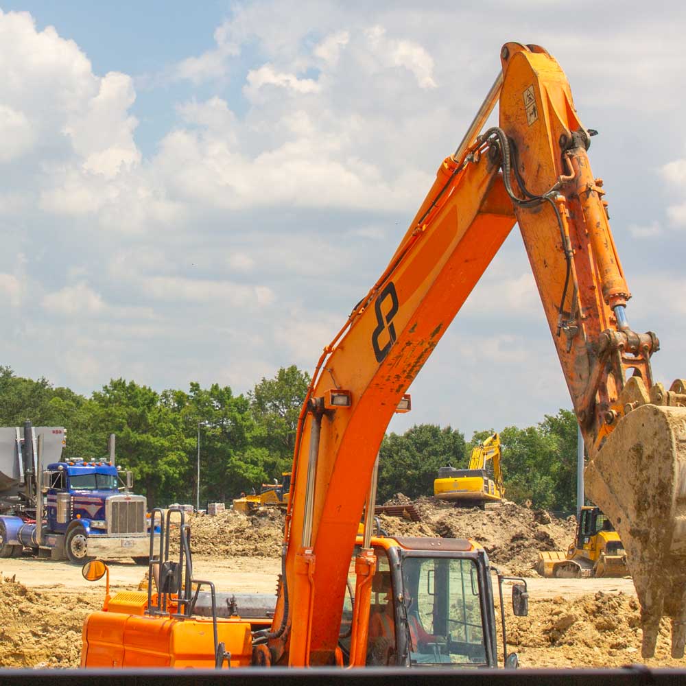 Various pieces of construction equipment clear out a space by digging up large mounds of dirt as a truck weaves between them, hauling out another load of that dirt. In the background stands a row of trees.