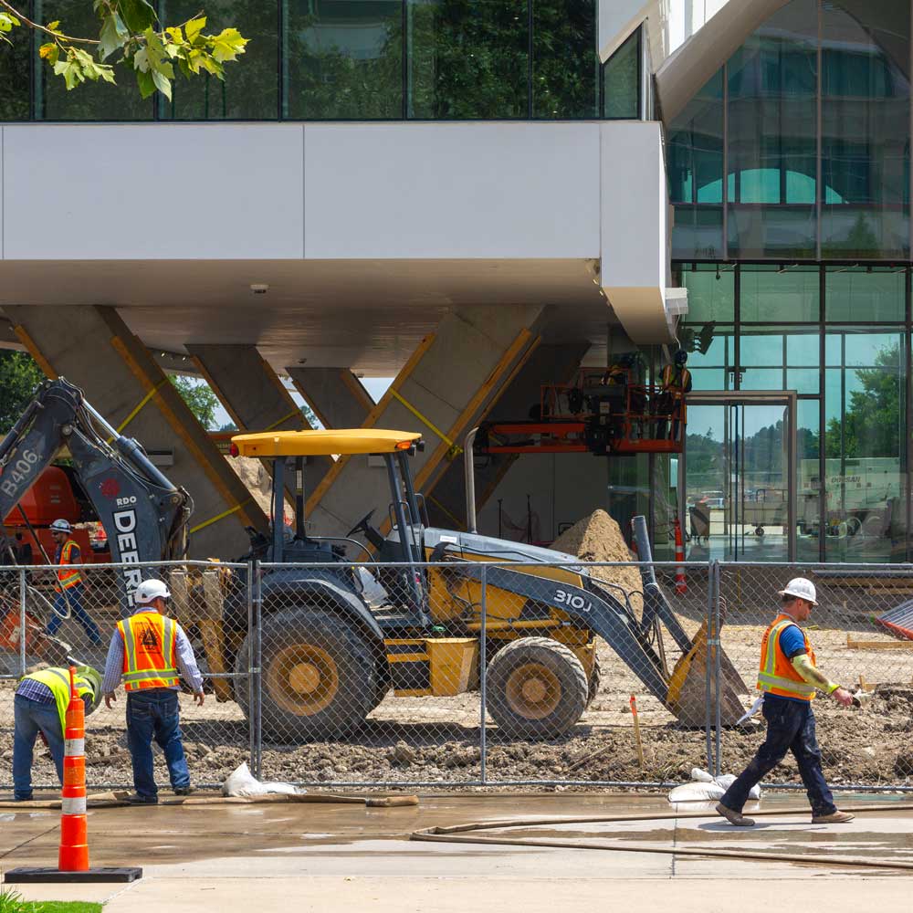 Construction workers and equipment move around below an elevated section of a modern glass, steel, and stone building supported on v-shaped columns.