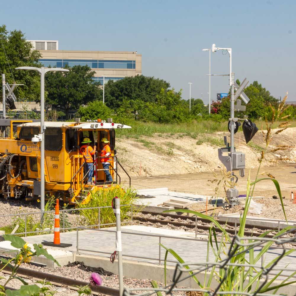 A train-like machine rolls across some recently-placed railroad tracks at the construction site for a train station and street intersection.