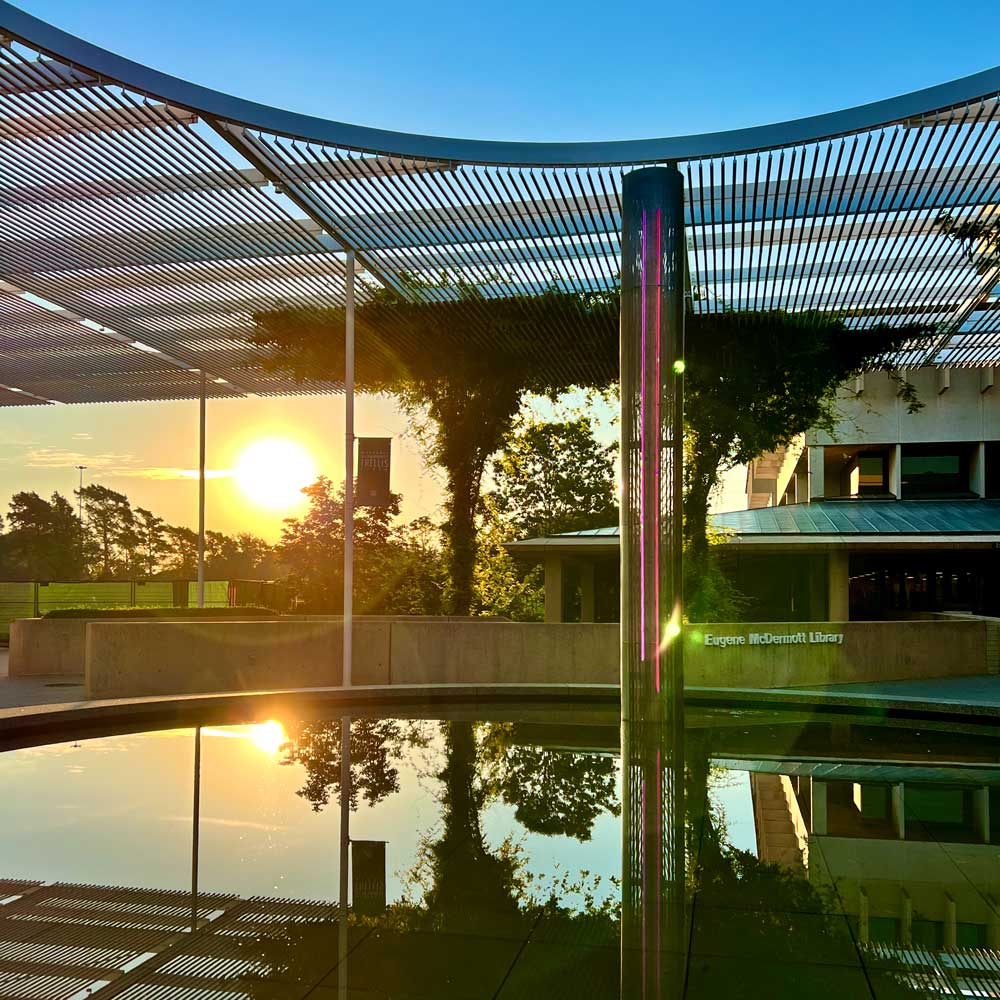 The morning sun rises between the a construction site and a brutalist-style building to shine in the water of a circular pool, from which rises a modern metal mesh and neon fountain. The pool rests just under a circular hole in a modern metal trellis that spans an entire plaza.