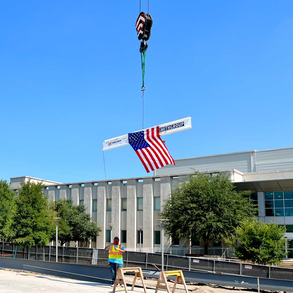 A crane raises a white I-beam, covered in signatures and trailing a US flag, high above a construction site.