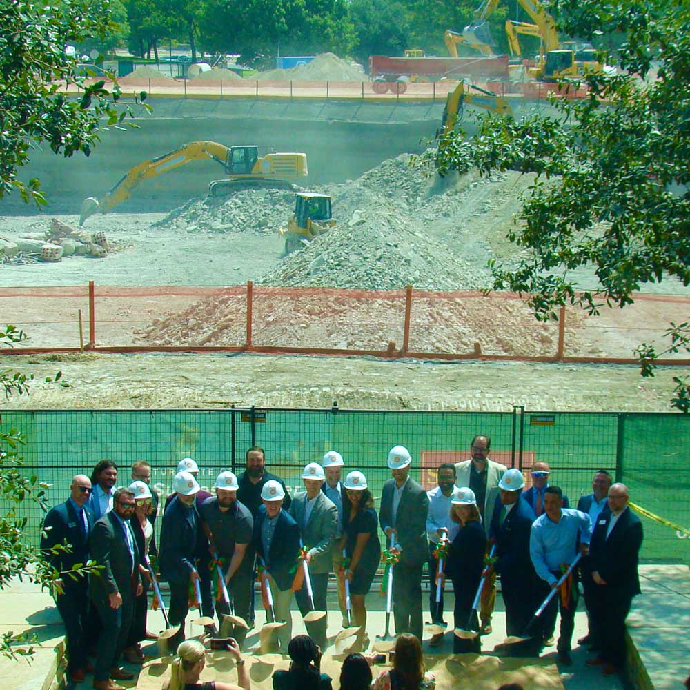 A group of officials with hardhats and shovels scoop dirt in front of a construction site as photographers take pictures of the ceremony.