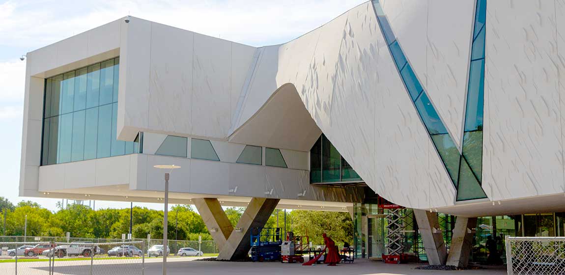 Facilities & Economic Development - The University of Texas at Dallas. The Edith and Peter O’Donnell Jr. Athenaeum. A modern building still under construction, formed from abstract shapes in textured stone and tinted glass, and raised in the air on v-shaped pillars. Under the building, various chery-pickers and tool carts are lined up next to a red human-sized sculpture of a monk with a broom.