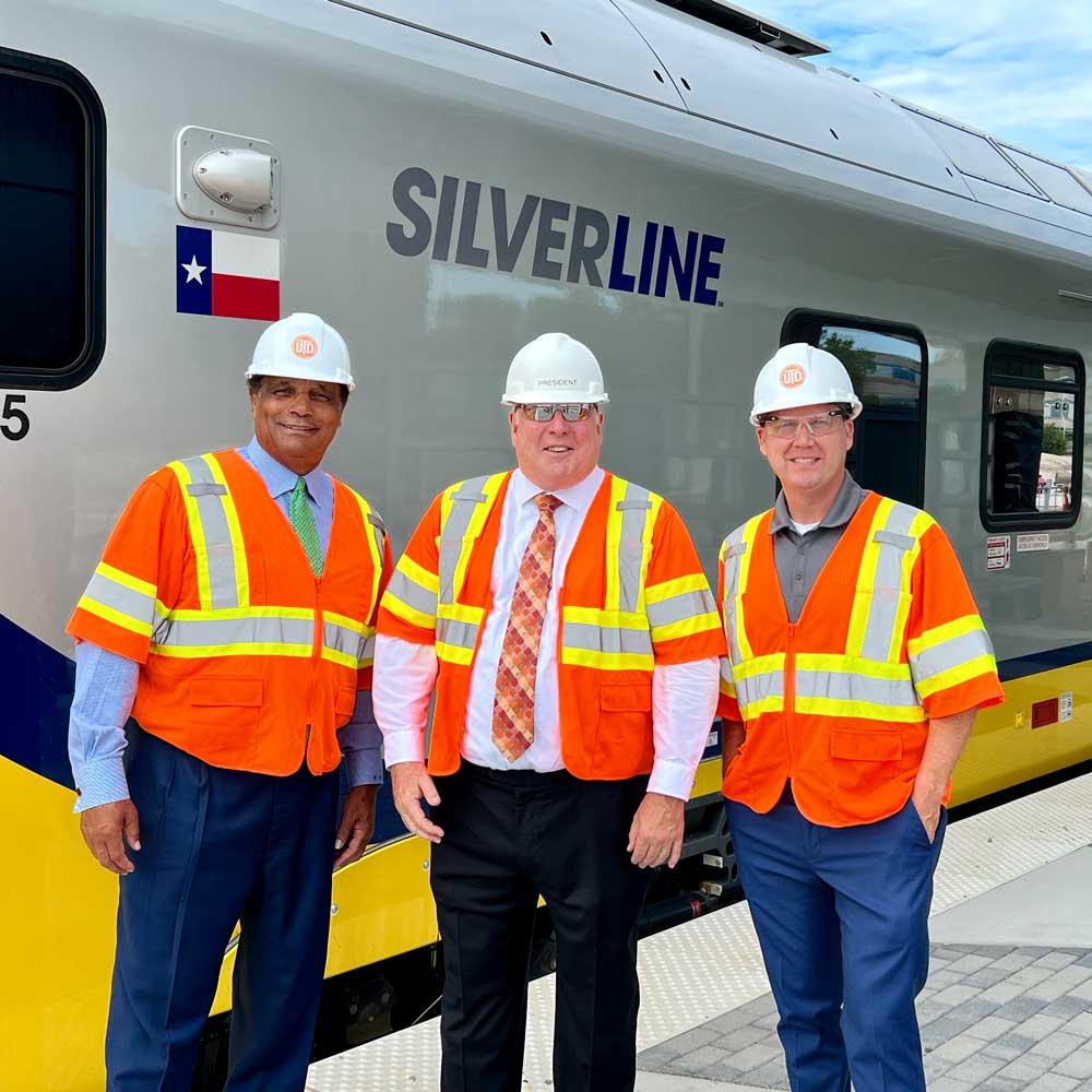 Three men in hardhats and safety vests stand in front of a new commuter train at a rail station construction site.