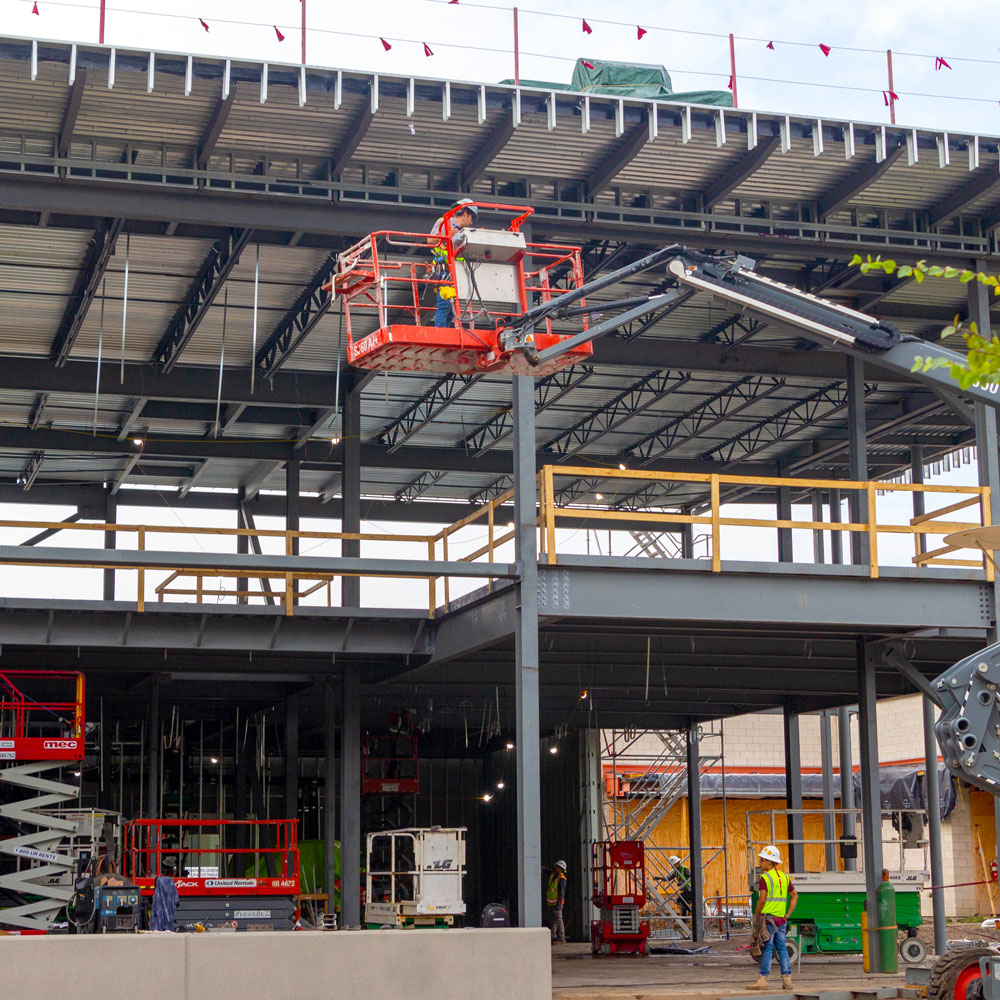 A construction worker rides a cherry-picker to the underside of a metal roof that extends over the framework of a two-story building.