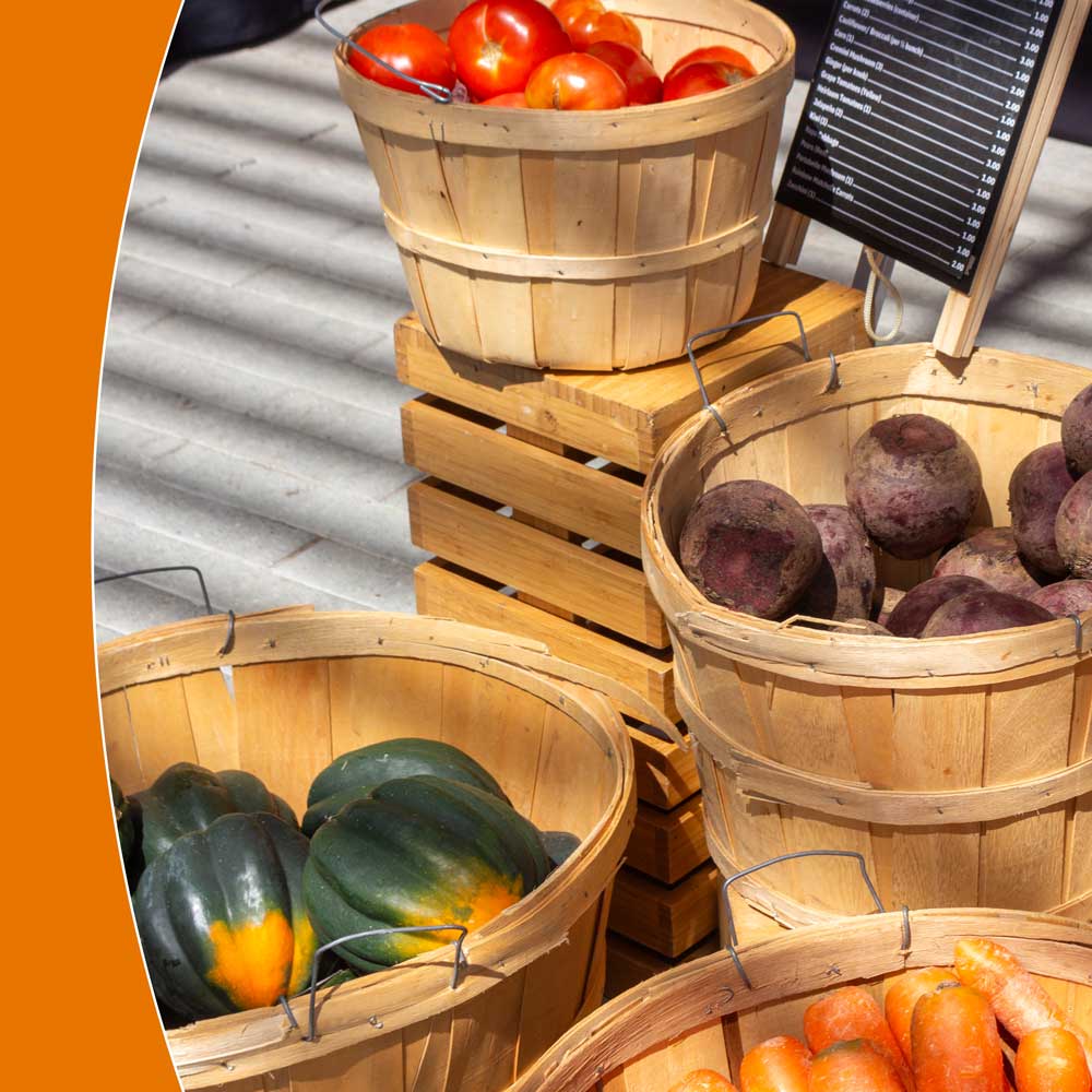 Baskets of fresh vegetables arranged on the ground and stacked atop crates near a small wooden sanwichboard posting their prices on a chalkboard.