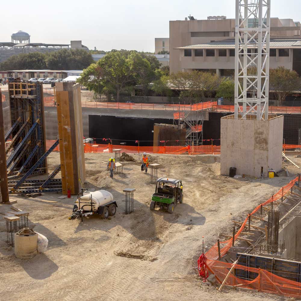 A construction site in the middle of a college campus where both a deep hole is being dug and high walls are being raised.