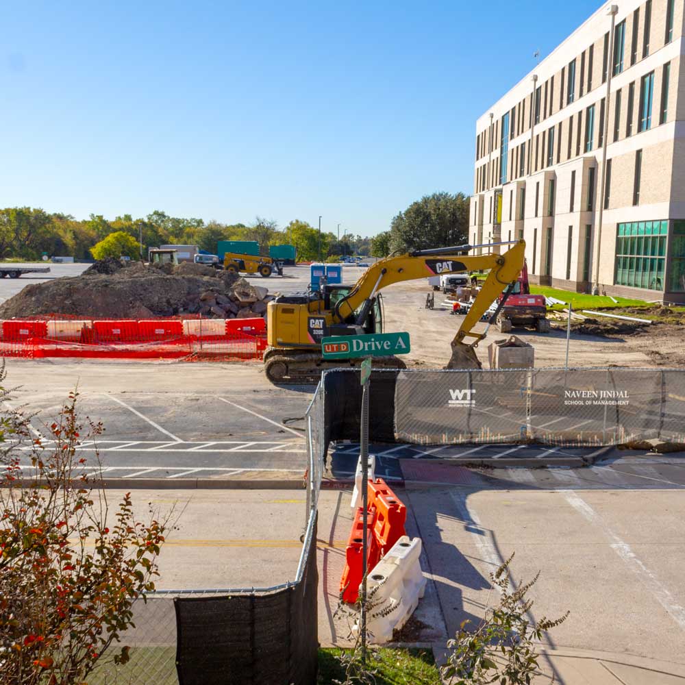 A construction excavator and other pieces of heavy construction equipment sit by freshly-made mounds of dirt in a parking lot next to a tan stone and brick building behind a construction fence. Text on the fence reads “Naveen Jindal School of Management”.