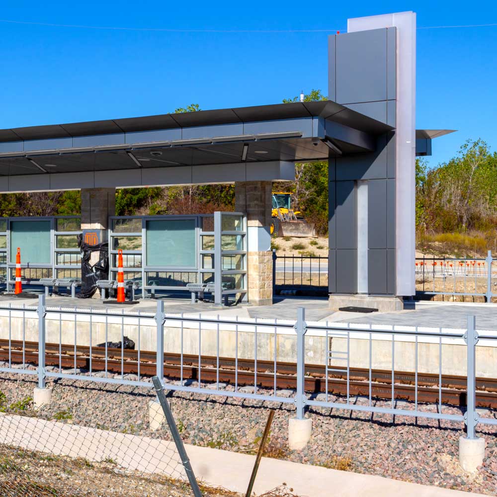 An under-construction train station in steel, glass, and stone. Some components are wrapped in black plastic, but one of those coverings has come free just enough to show the UTD mongram underneath.