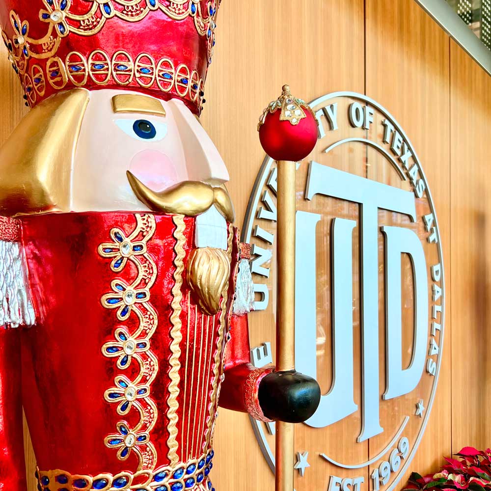 A giant replica of an ornamental nutcracker in reds and golds stands against a wooden wall bearing the logo for the University of Texas at Dallas.