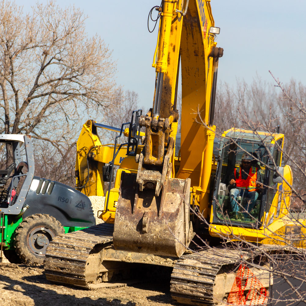 A trio of construction machines poised on a patch of bare soil between leafless trees.