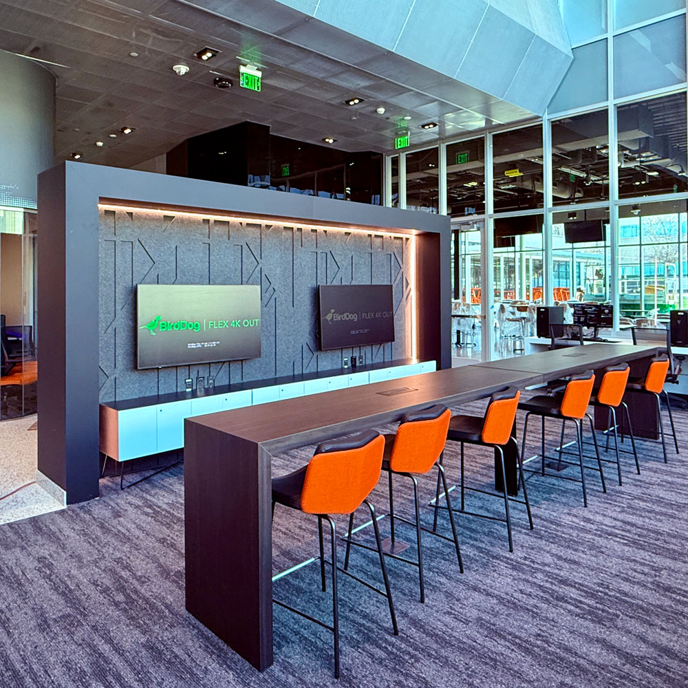 A portion of a modern lounge rendered in glass, mirrored and textured metal, and polished wood. Several raised orange chairs are tucked into a long table in front of a pair of large monitors. To both sides are hints of larger spaces beyond the photo’s frame.