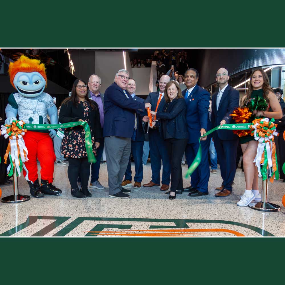 A group of people lined up behind a ribbon, just inside the entrance to a modern steel, glass and stone facility, with the stylized letters “UTD” decorating the floor. The ribbon has just been cut and is fluttering to the ground.