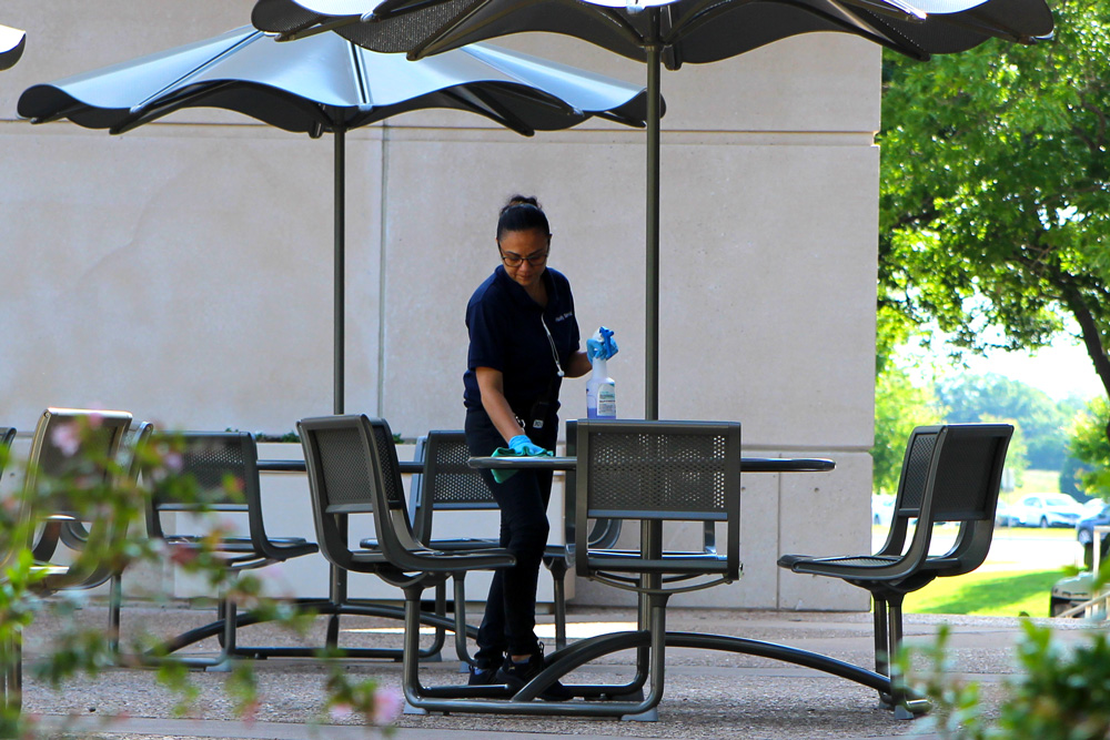 A custodial worker cleaning umbrella-coverd tables outdoors.