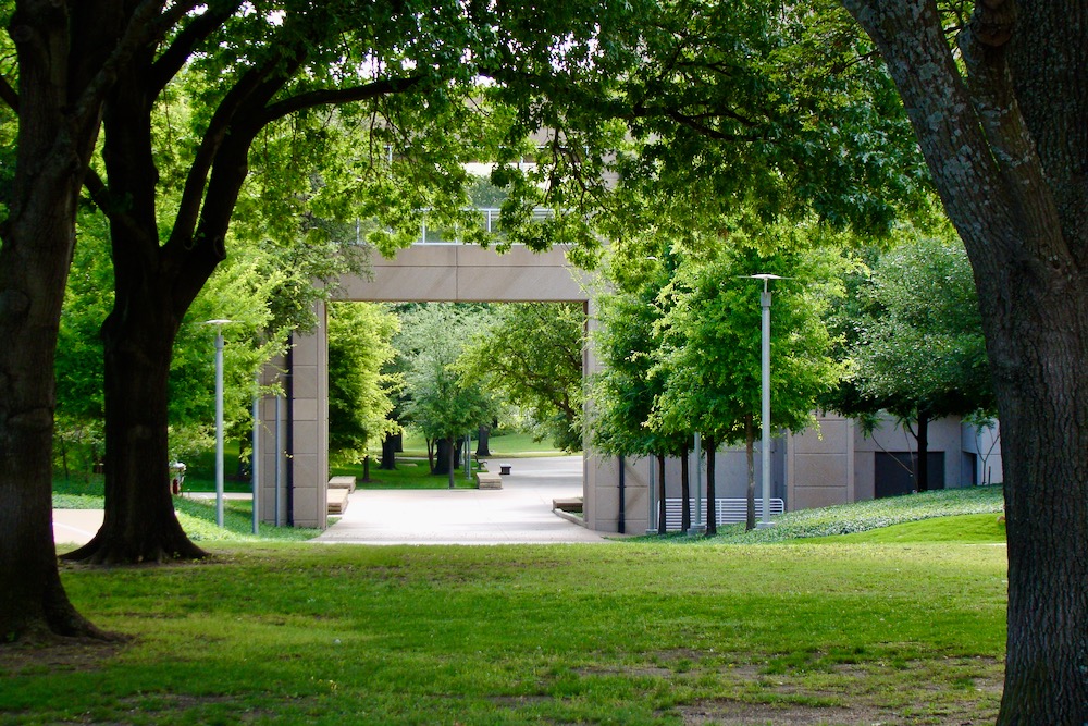 The tree canopy creates a natural arch which frames a concrete and glass arch formed by the skywalk connecting the Administration Building to the Erik Jonsson Academic Center.