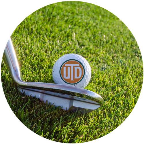 A golf ball bearing the UTD monogram sits on the grass of a golf course, about to be hit by a golf club. Above it are the words “Greens for Dreams”.